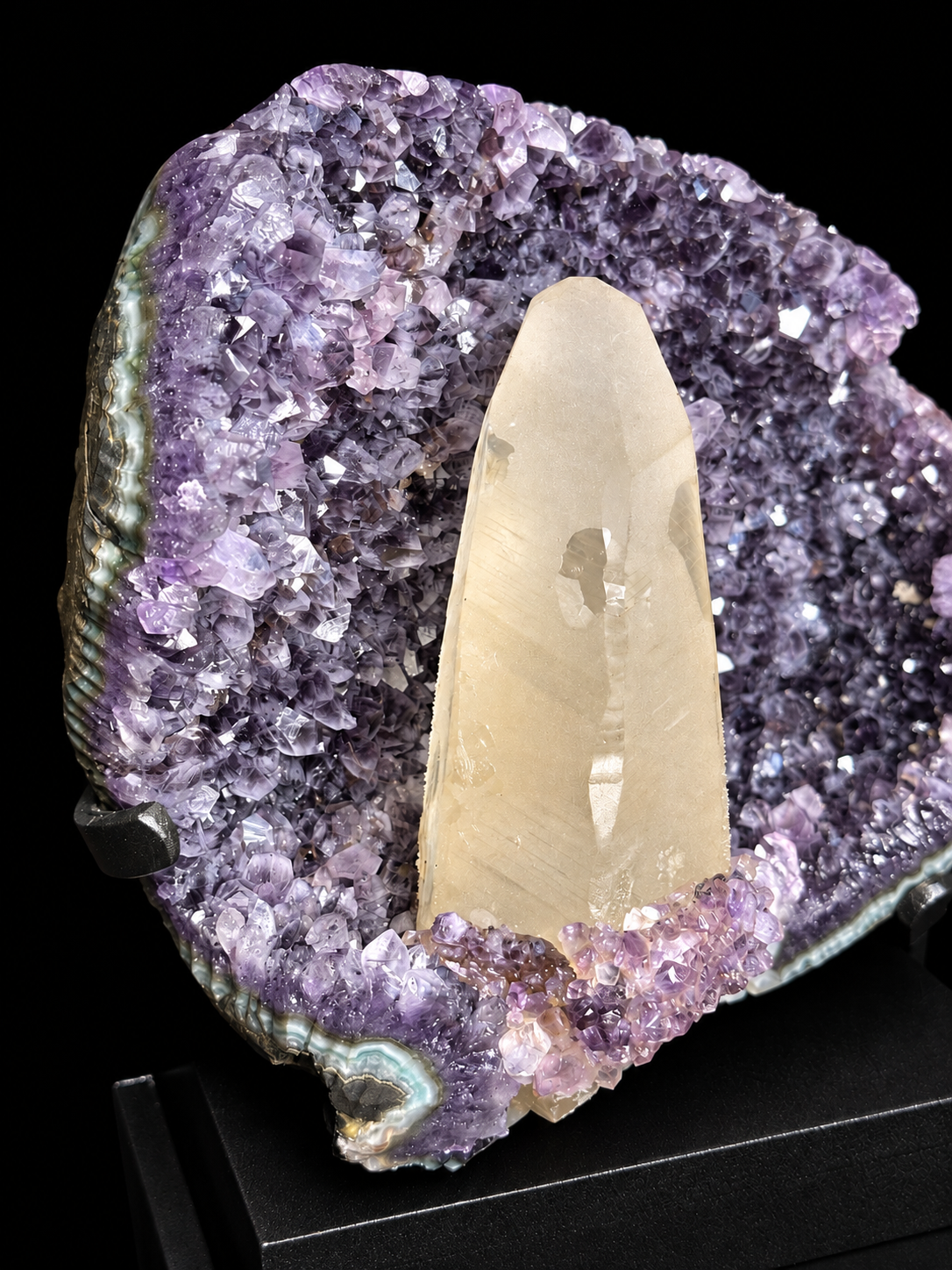 A natural amethyst geode display with a prominent ivory calcite crystal point set inside a purple amethyst cavity, mounted on a black display stand.