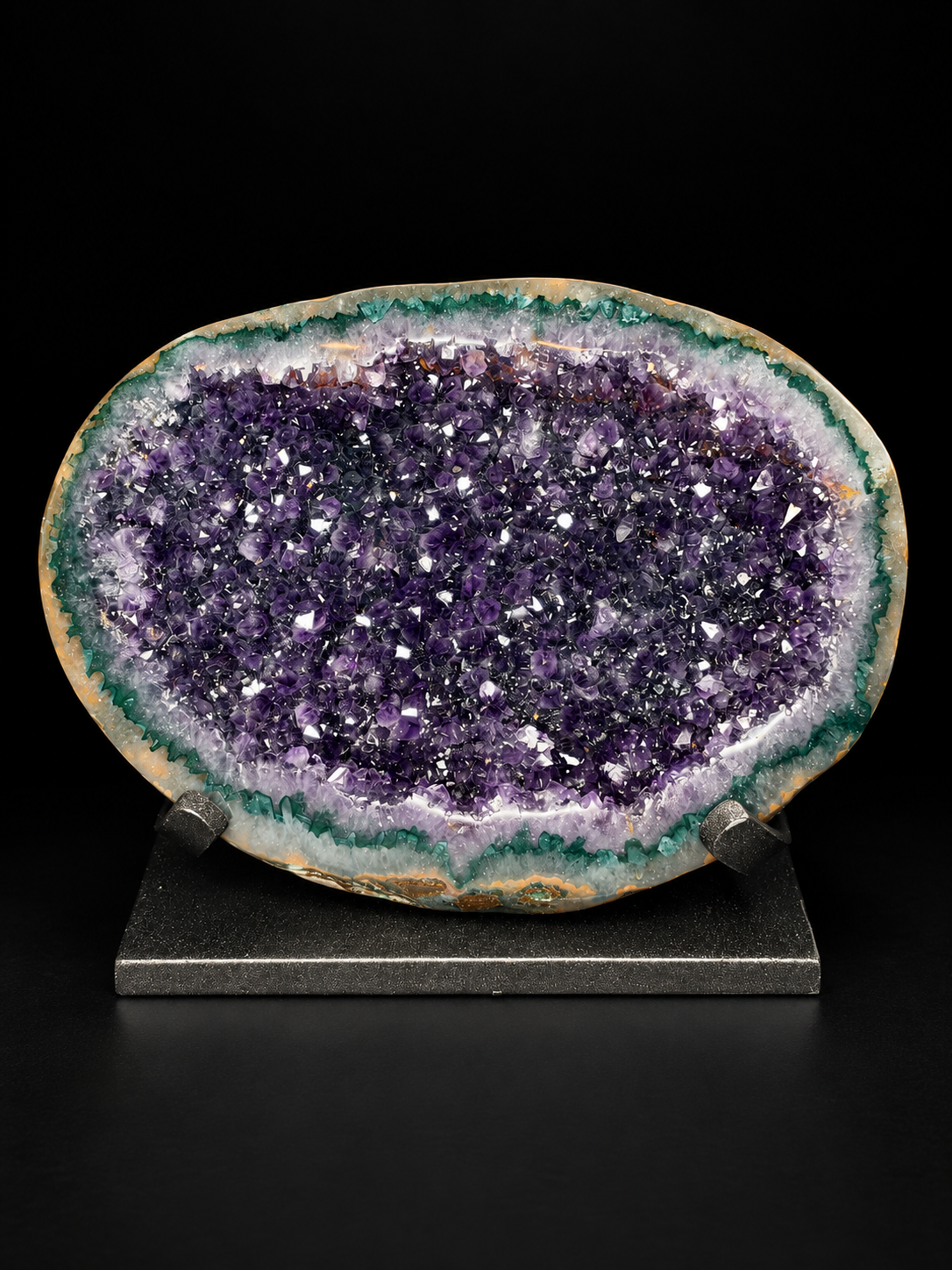 A polished amethyst geode specimen with a deep purple crystal interior, green and white outer matrix, and a natural irregular outline, displayed on a small dark stand against a black background.
