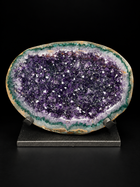 A polished amethyst geode specimen with a deep purple crystal interior, green and white outer matrix, and a natural irregular outline, displayed on a small dark stand against a black background.