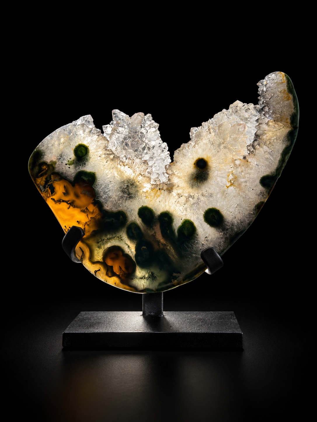 A polished agate geode slice with natural crystal clusters and dark green to amber inclusions, displayed on a black stand and backlit.