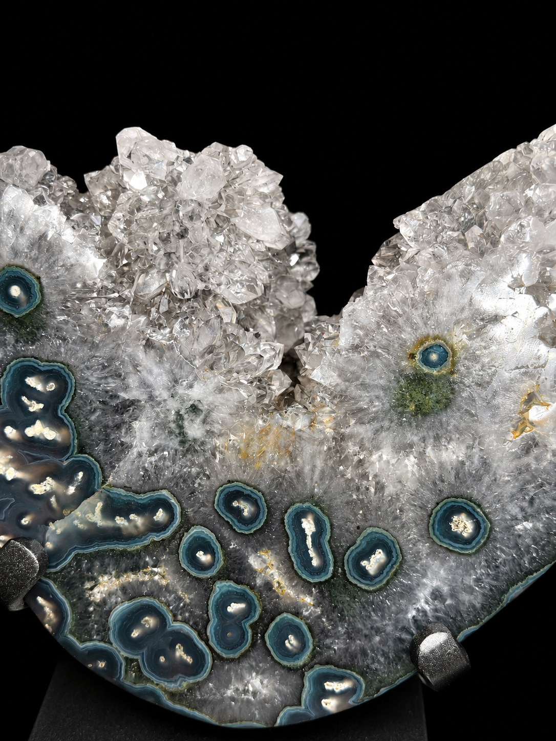A geode specimen with clear quartz crystal clusters and concentric blue-green stalactite "eyes" on a dark black base and background.