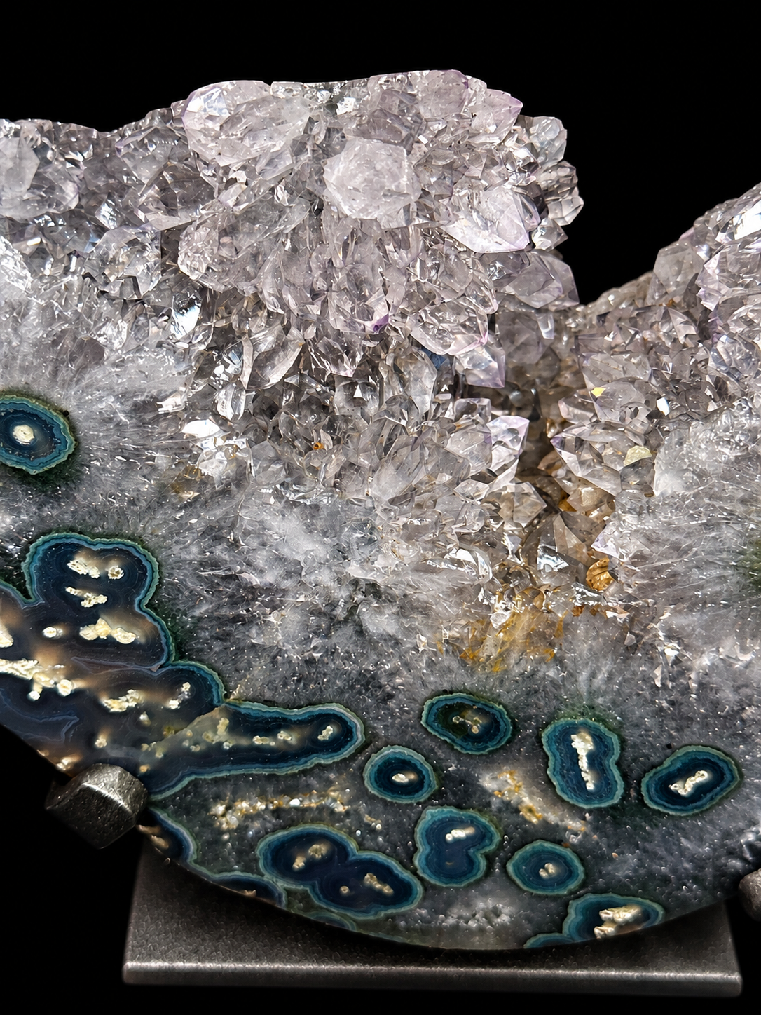 A close-up of a geode calcite and quartz mineral specimen with clear quartz crystals on top and concentric blue, teal, and tan agate bands, mounted on a black display stand against a black background.