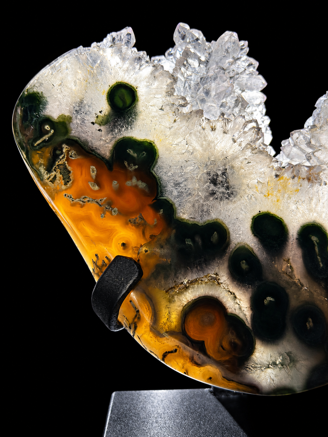 A polished agate stone specimen with a natural crystal cluster on a black background, mounted on a display stand. The stone shows amber, dark green, black, and clear crystalline patterns when backlit.