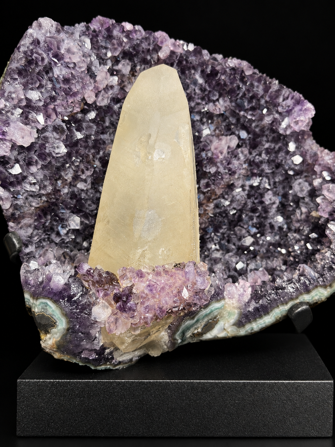 A natural amethyst geode display with a prominent ivory calcite crystal point set inside a purple amethyst cavity, mounted on a black display stand.