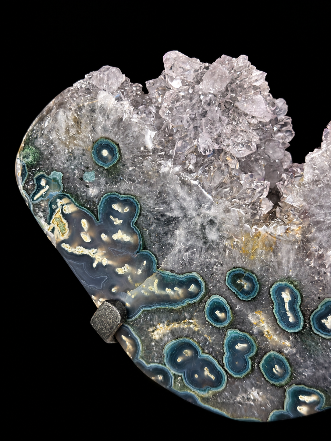 A polished agate geode specimen with concentric blue-green bands and topped with clear quartz crystals, mounted on a black base against a solid black background.