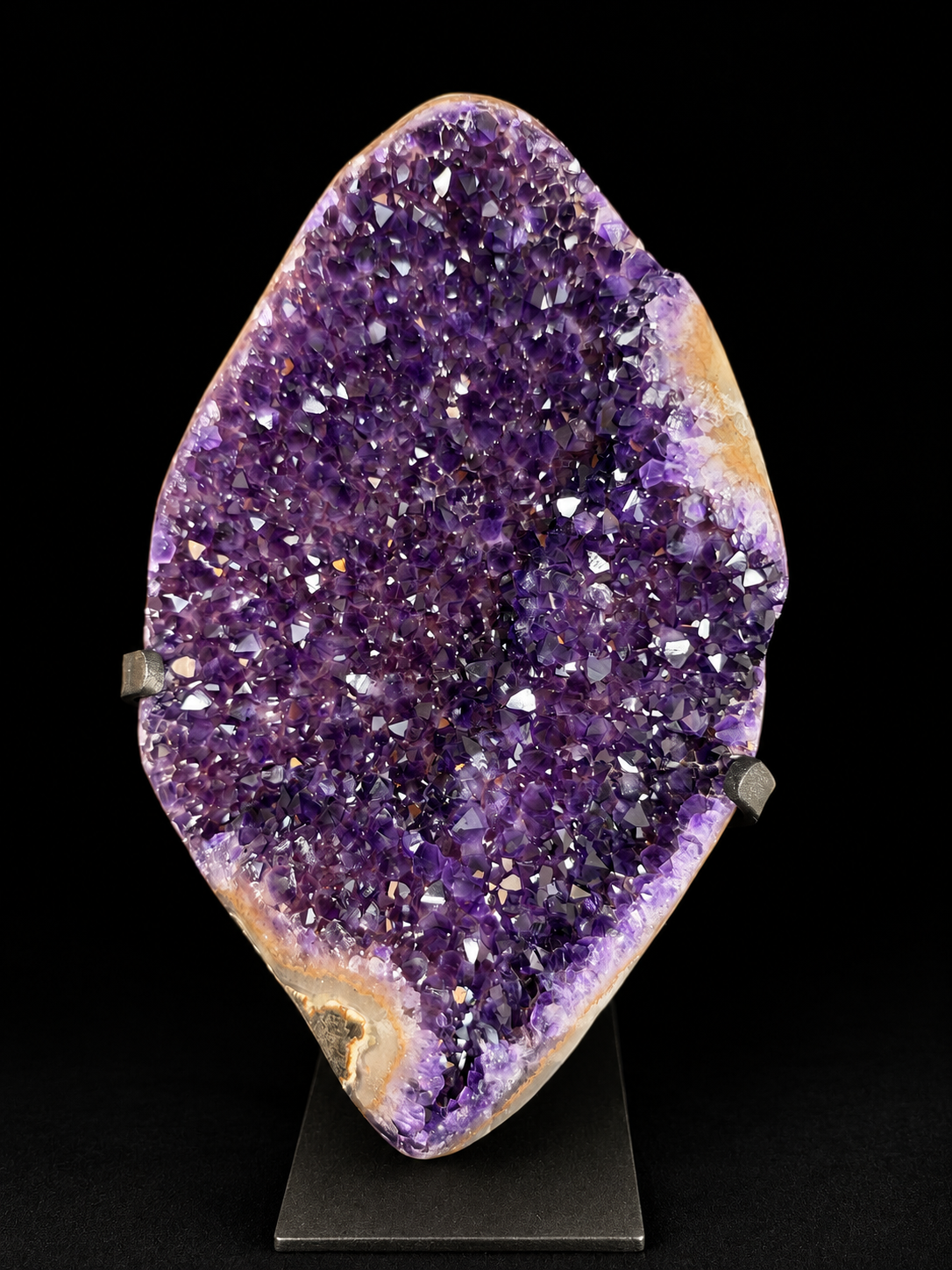 A large natural amethyst geode specimen with a deep purple crystal interior and tan outer matrix, displayed upright on a black stand against a black background.