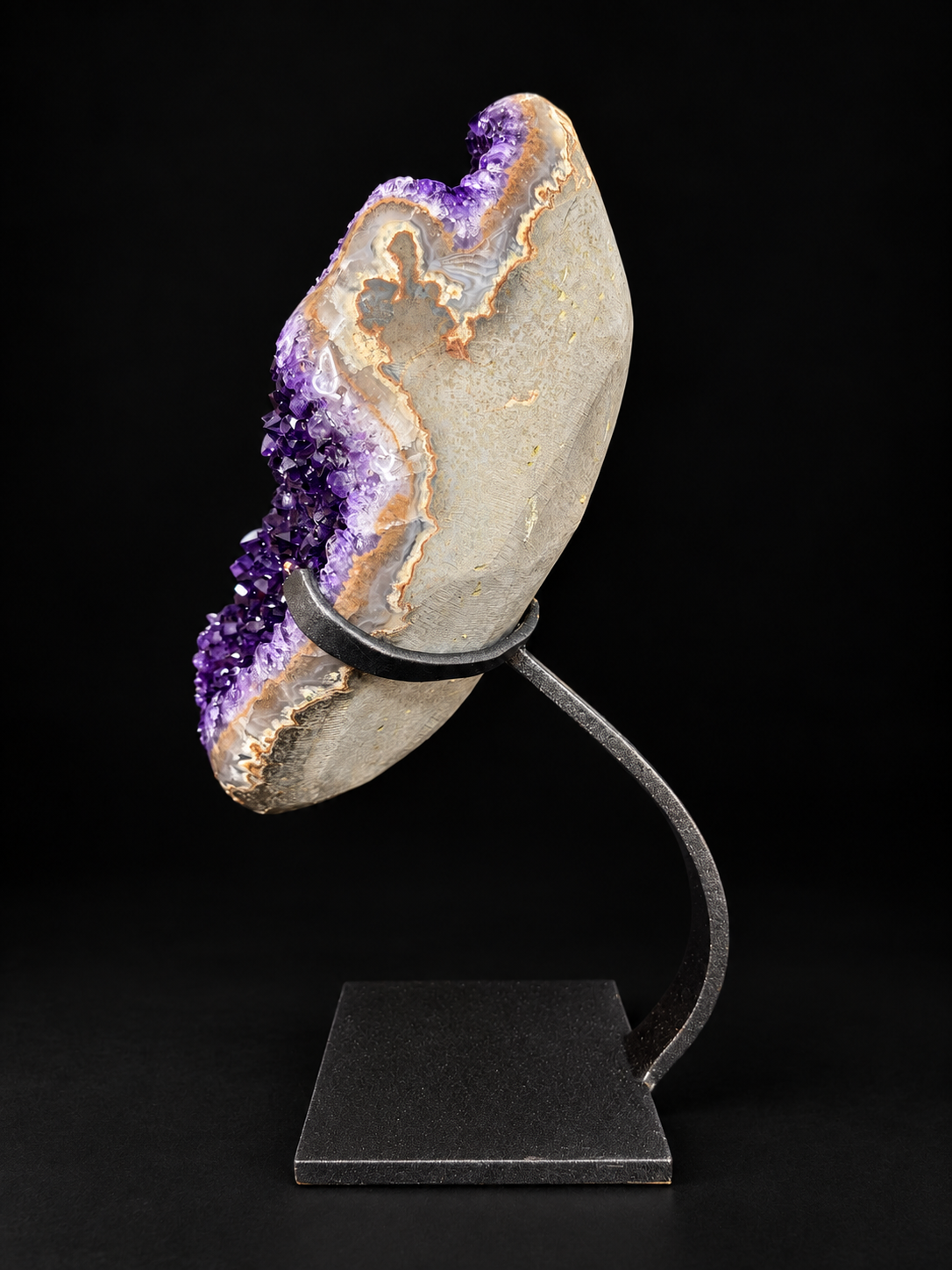 A natural amethyst geode specimen mounted on a black metal display stand against a solid black background.