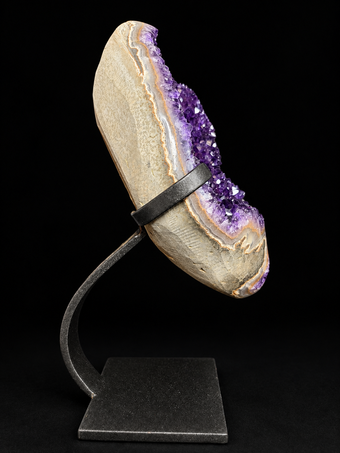 A raw amethyst geode display piece mounted on a black metal stand against a solid black background.