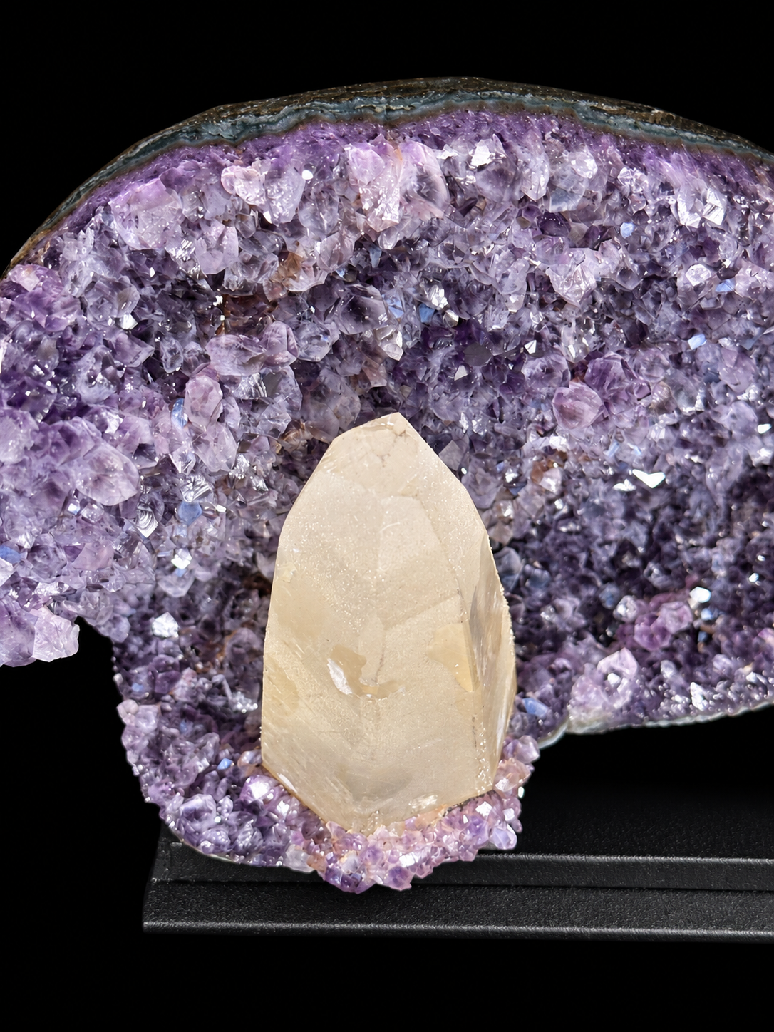 A natural amethyst geode display with a prominent ivory calcite crystal point set inside a purple amethyst cavity, mounted on a black display stand.